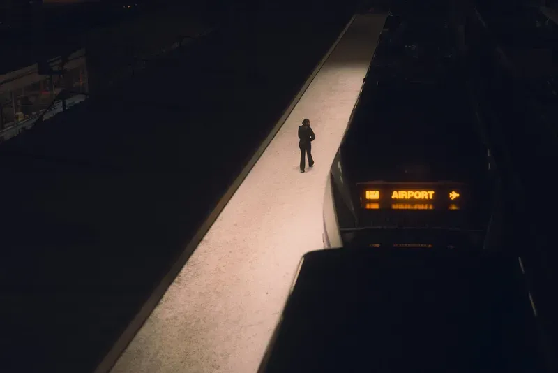 Seen from above, a figure in dark clothing walks alone along a lit elevated walkway toward a train marked Airport at night.
