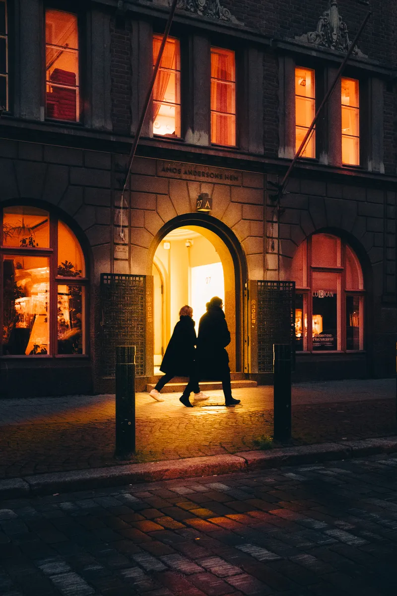 Two people walk through a glowing stone archway into the Amos Andersons hem building at night, warm light spilling onto the cobblestones.