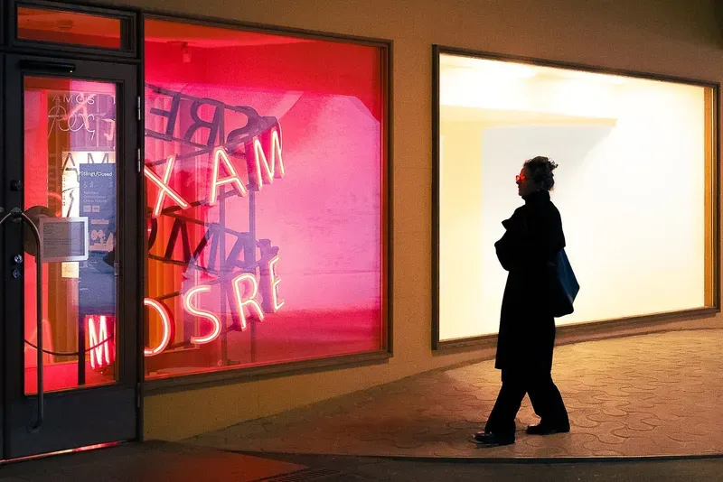 A mirrored red neon sign reading Amos Rex glows in a window as a woman in dark sunglasses and a long coat walks past a brightly lit gallery storefront at night.