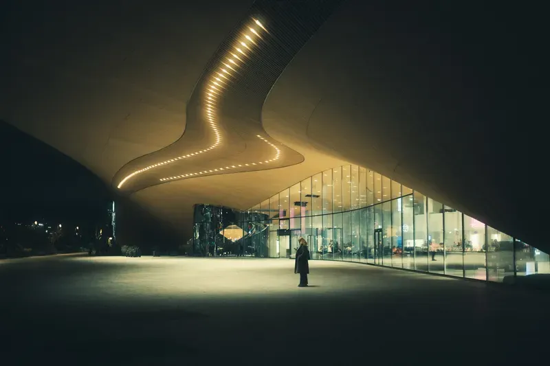 A lone figure checks their phone beneath the curved, glowing ceiling ribs of Amos Rex museum's plaza at night, its glass facade lit from within.