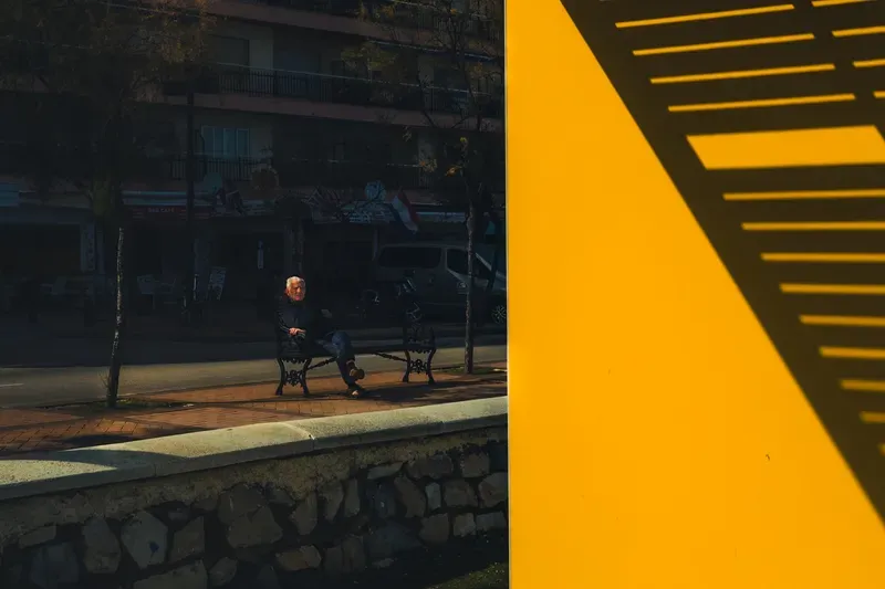 An elderly man sits alone on a park bench across a street lined with flags and cafes, seen past the sharp diagonal edge of a sunlit orange sign.