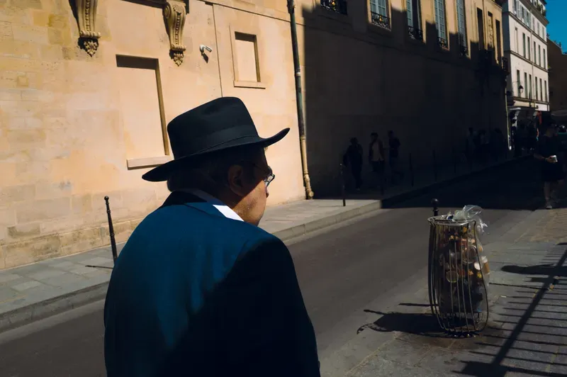 An older man in a black wide-brimmed hat and blue jacket looks down a sunlit street lined with pale stone buildings.