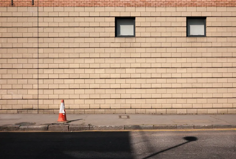 A dark cloud-shaped smoke stain marks a sunlit tan brick wall between two small windows above an empty sidewalk.