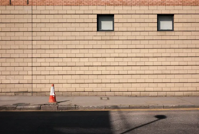 A dark cloud-shaped smoke stain marks a sunlit tan brick wall between two small windows above an empty sidewalk.