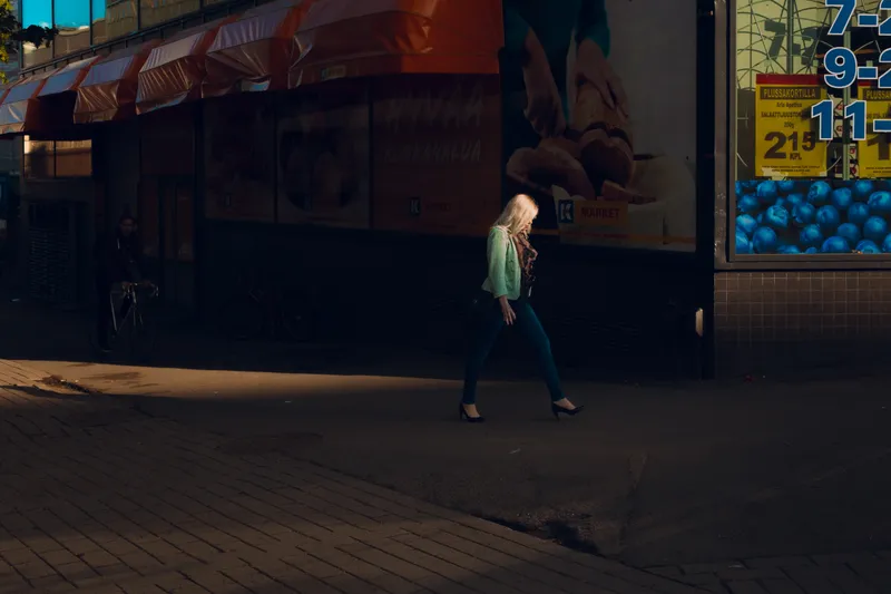 A blonde woman in heels crosses the street past a lit market storefront at night, a cyclist passing behind her.