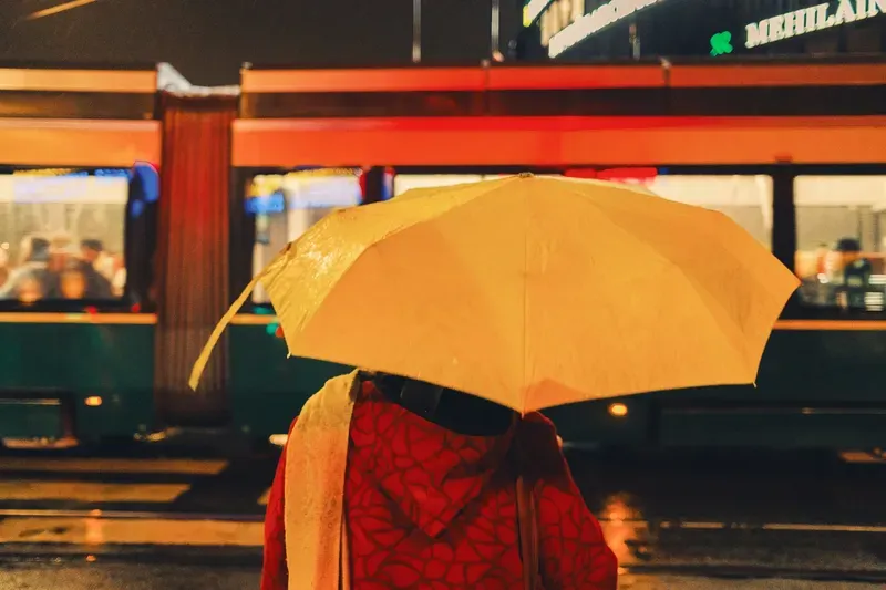A man in a yellow jacket and red trousers stands on a train platform at blue hour, breath visible in the cold air between passing trains.