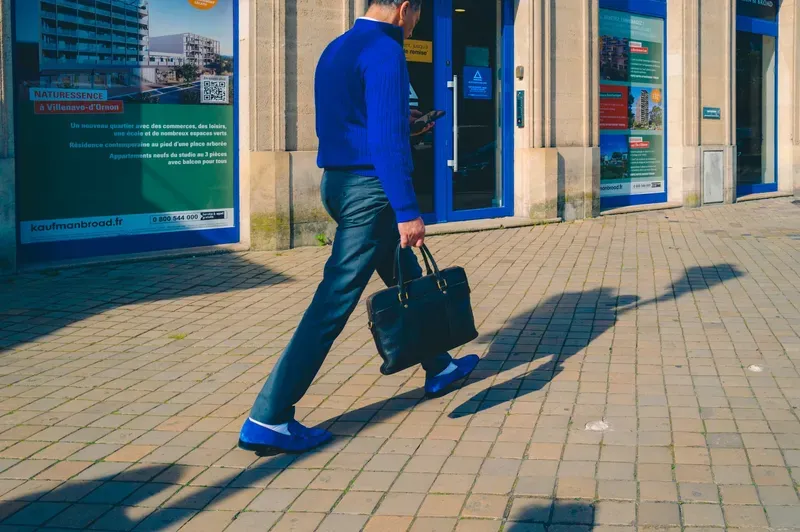 A man in a blue sweater and suede loafers checks his phone while carrying a leather bag past a real estate storefront.