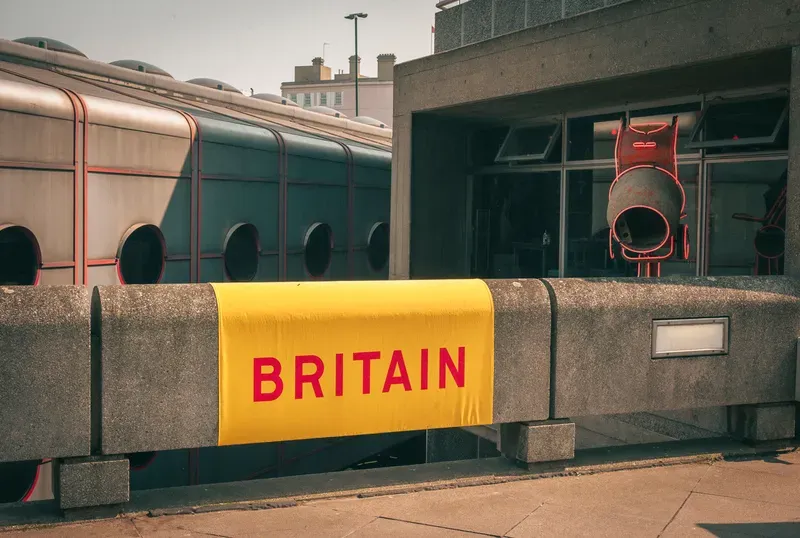 A yellow 'Britain' banner drapes over a concrete barrier in front of a brutalist building with a suspended cement mixer sculpture outlined in neon.