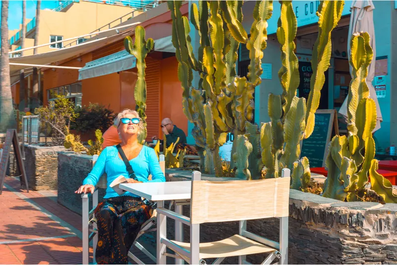A woman in sunglasses tilts her face to the sun at an outdoor café table surrounded by tall cactus plants.