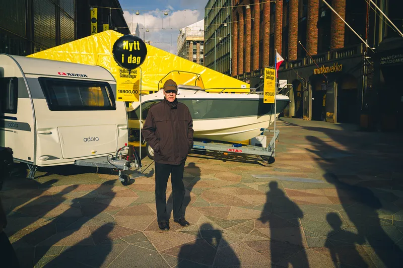 A man in a cap stands between a caravan and a boat on display for sale in a sunny plaza, surrounded by long shadows of onlookers.