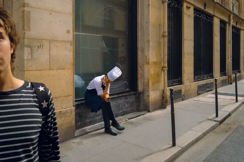 A chef in whites and a tall hat sits in a shop window recess looking at his phone, while a young man in a striped shirt passes close to the camera.