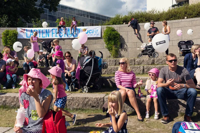 Families with balloons and pink sunhats gather on stone steps at a children's festival, kids perched on the wall above a banner.
