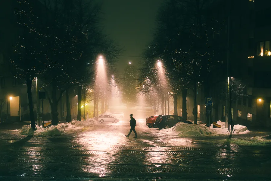 A person crosses a street lined with Christmas-lit trees, wet cobblestones reflecting the lights.