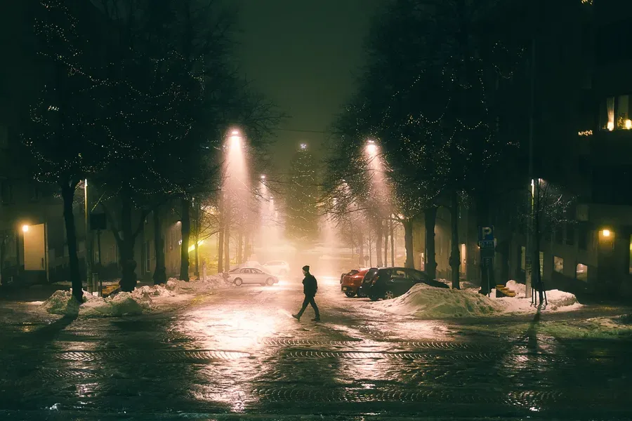A person crosses a street lined with Christmas-lit trees, wet cobblestones reflecting the lights.