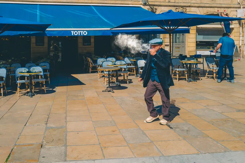 A man in a flat cap exhales a cloud of smoke while walking past an empty cafe terrace with blue umbrellas and striped chairs.