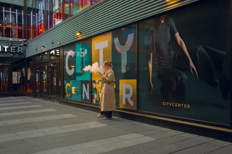 A woman in a trench coat exhales a white cloud of vapour that hides her face, standing at night against the teal and gold lettering of a lit shopping centre hoarding.