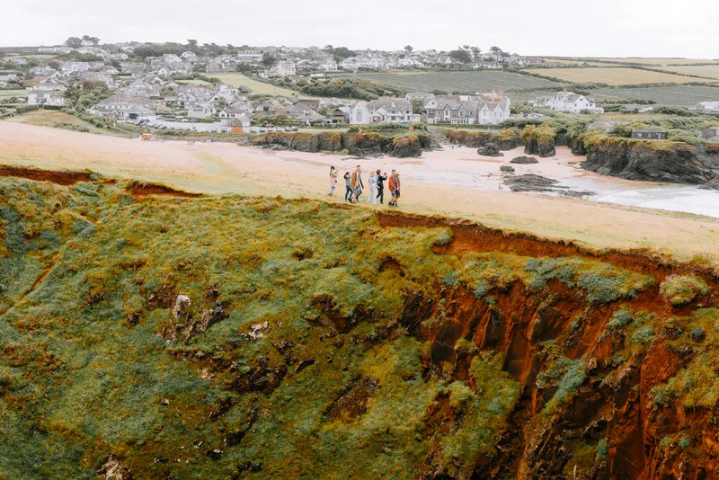 Seen from above, a small group walks along a grassy cliff edge overlooking a sandy beach and a seaside town.