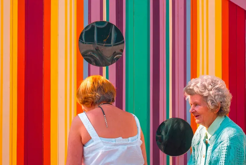 An elderly woman in a green cardigan talks beside a younger woman, both standing before a wall of rainbow-striped panels with circular mirrors.