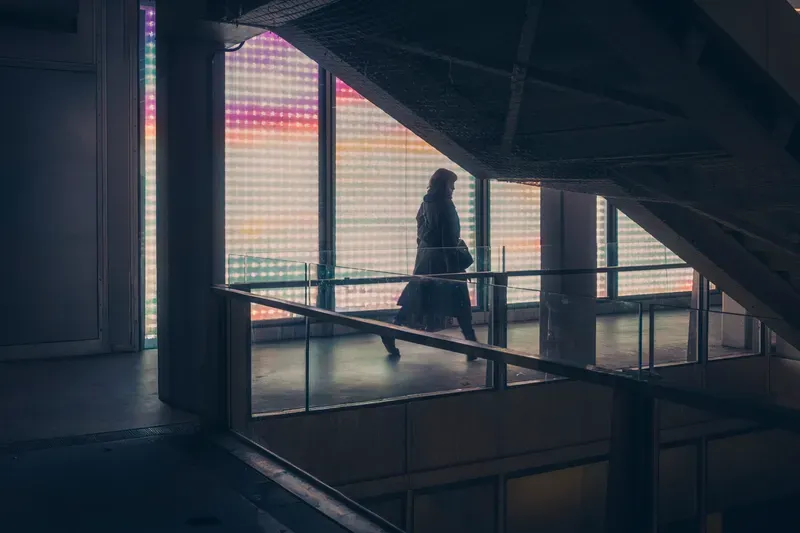 A woman in a long coat is silhouetted against a rainbow-lit glass wall as she crosses a glazed walkway.