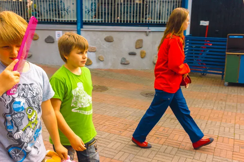 A boy raises a pink comb in front of his face beside a boy in a bright green t-shirt as a girl in a red top and blue trousers strides past on a brick-paved street.