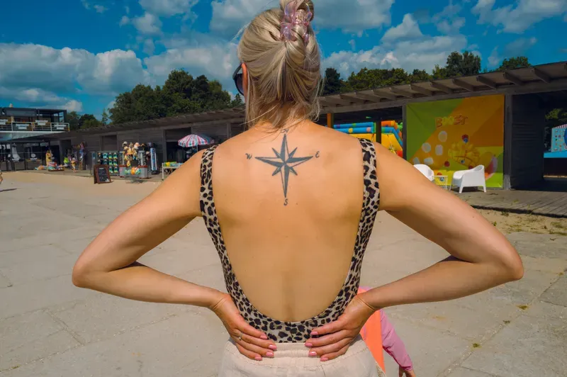 Seen from behind, a woman in a leopard-print swimsuit stands with her hands on her hips, a compass rose tattooed between her shoulders, facing a row of beach kiosks.