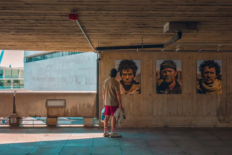 A man in red shorts pauses in a concrete underpass lined with large portrait photographs on the wall.