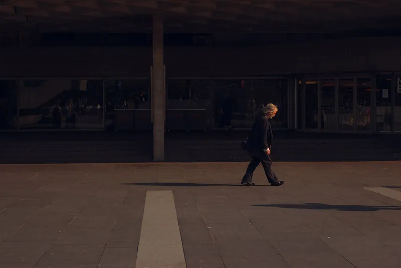 A woman in dark clothing walks alone across a shadowed concrete plaza, sunlight catching her hair.
