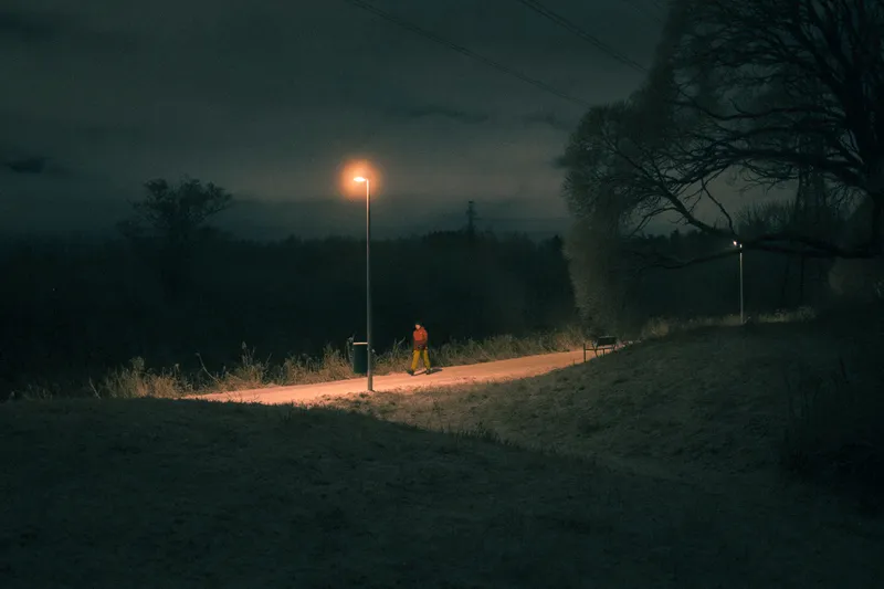A lone figure in a bright orange jacket walks a path lit by a single streetlamp, dark forest silhouetted against the night sky.