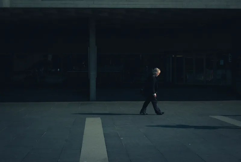 A lone figure crosses a dim, near-empty plaza beneath a concrete overhang.