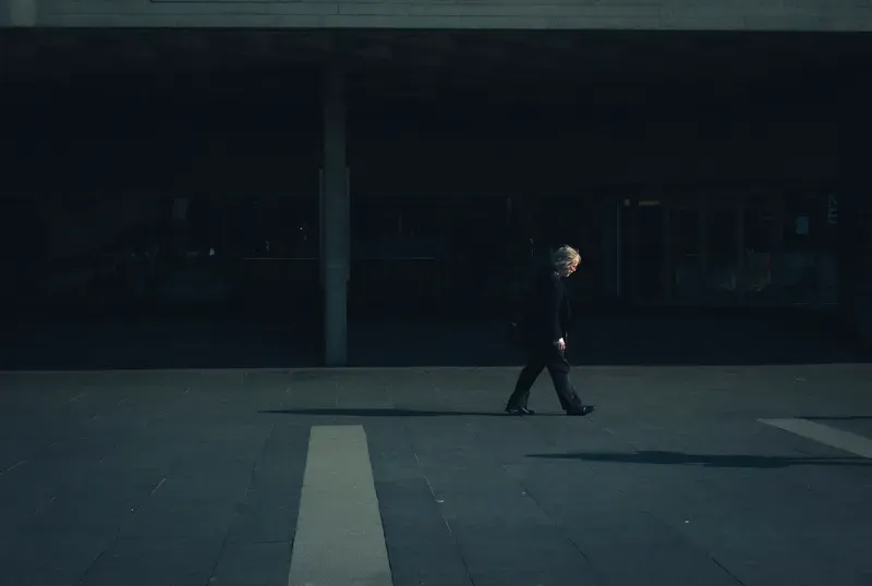 A lone figure crosses a dim, near-empty plaza beneath a concrete overhang.