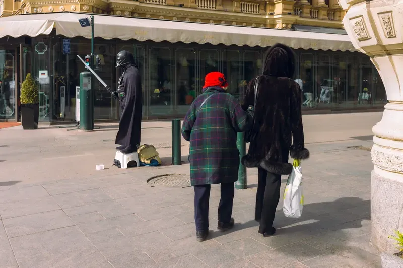 Two women seen from behind, one in a red hat and tartan coat, the other in a fur-trimmed coat, walk arm in arm past a street performer dressed as Darth Vader standing on a stool with a lightsaber.