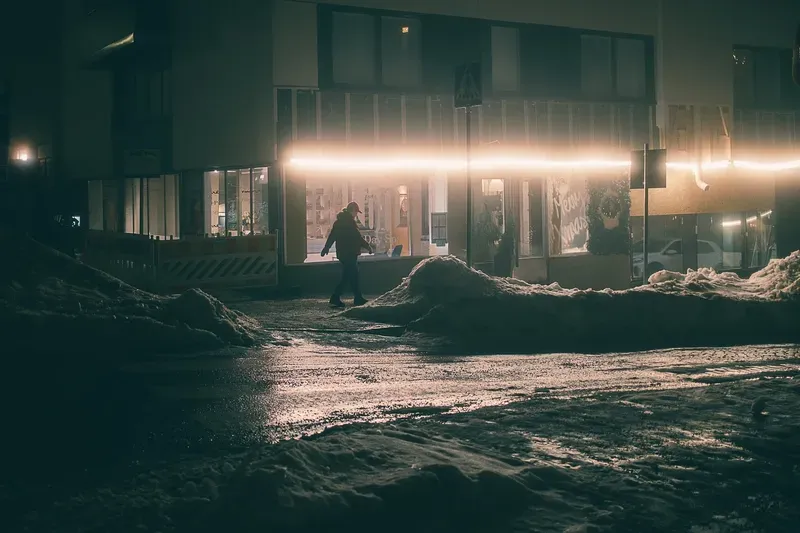 A person walks past an illuminated shopfront and a glowing neon light strip on a snowy commercial street at night.