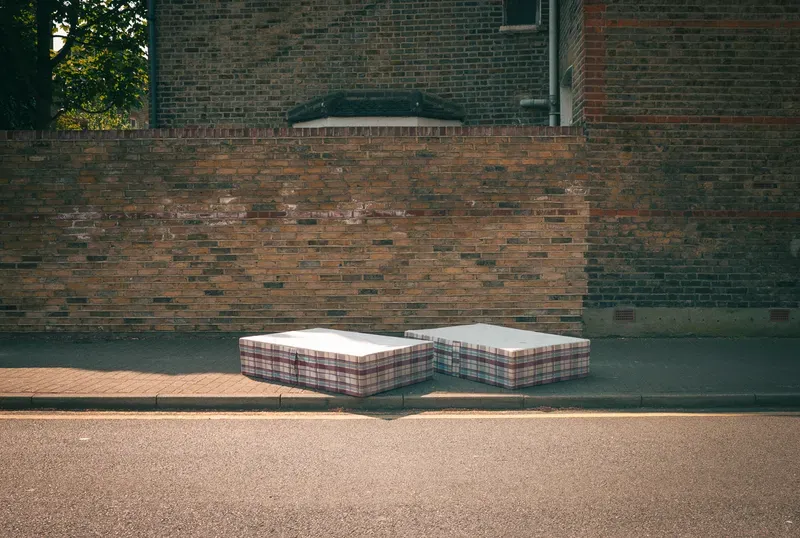 Two plaid mattresses lean against a curb on a sunlit sidewalk beneath a weathered brick garden wall.