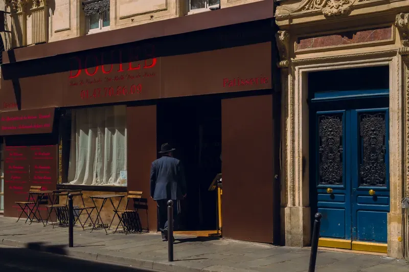 A man in a hat and dark suit steps through the open doorway of a deep red patisserie, beside a tall blue door with ornate ironwork.