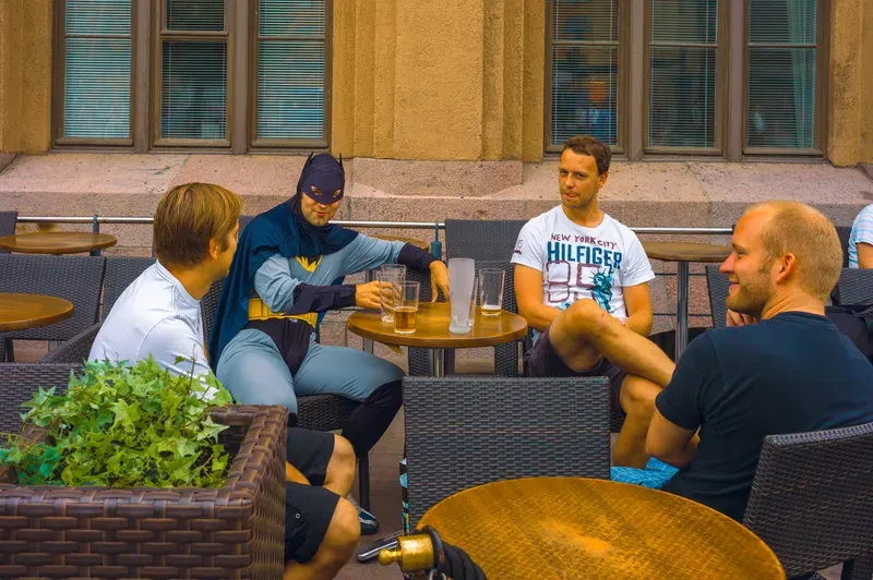 A man in a Batman costume sits at a café terrace table with three other men and glasses of beer, a stone building behind them.