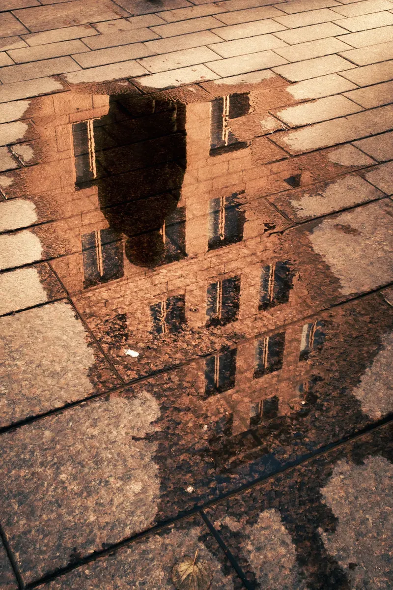 A building's upper stories and a passerby's silhouette are reflected in a puddle set into a warm-toned cobblestone street.