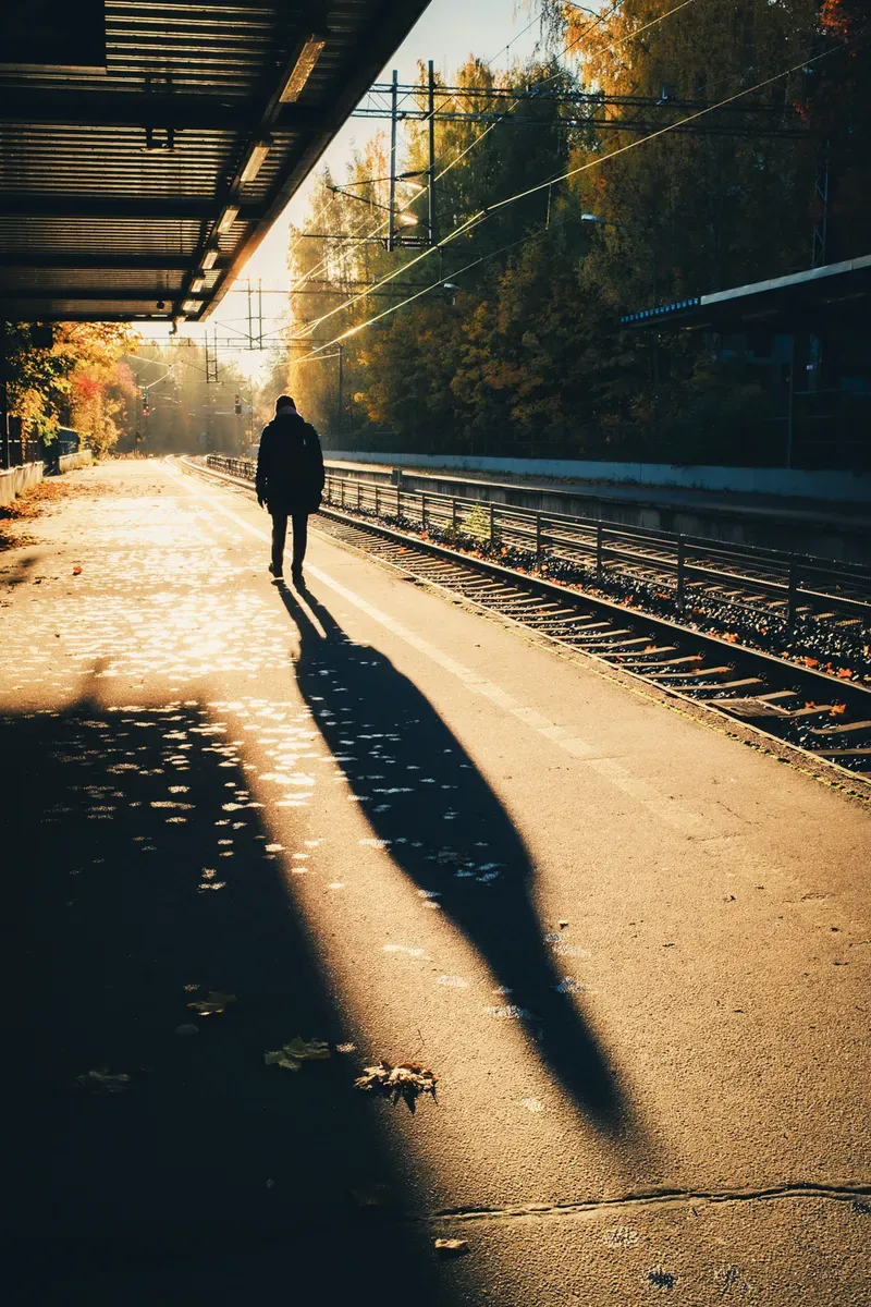 A silhouetted figure walks along railway tracks in low golden light, a long shadow trailing behind.