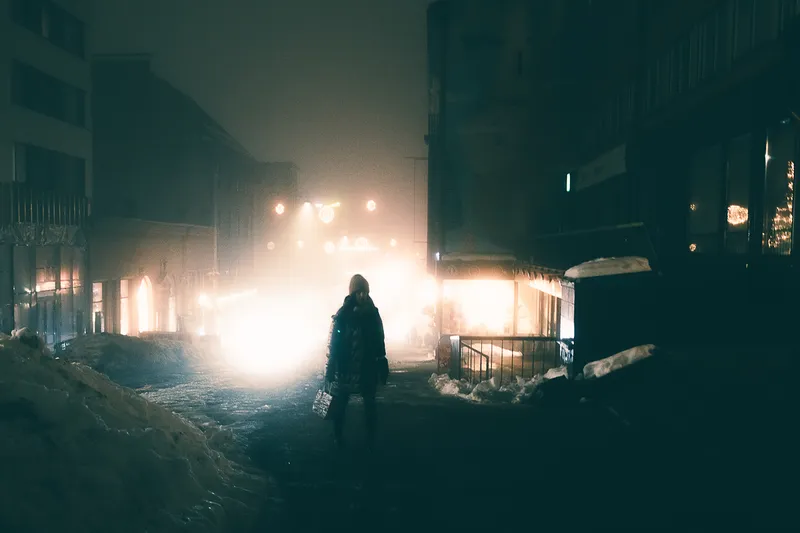 A bundled figure stands in a snow-piled street at night, streetlights and shopfronts diffused into a haze of fog and falling snow.