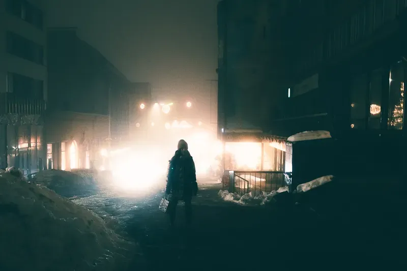 A bundled figure stands in a snow-piled street at night, streetlights and shopfronts diffused into a haze of fog and falling snow.