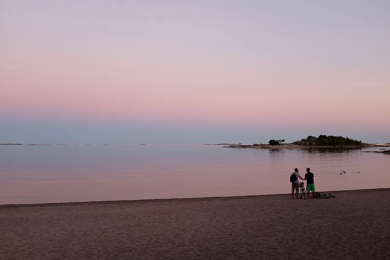 Two men and their bicycles stand silhouetted on a beach at dusk, the pink sky reflected perfectly across the still water.