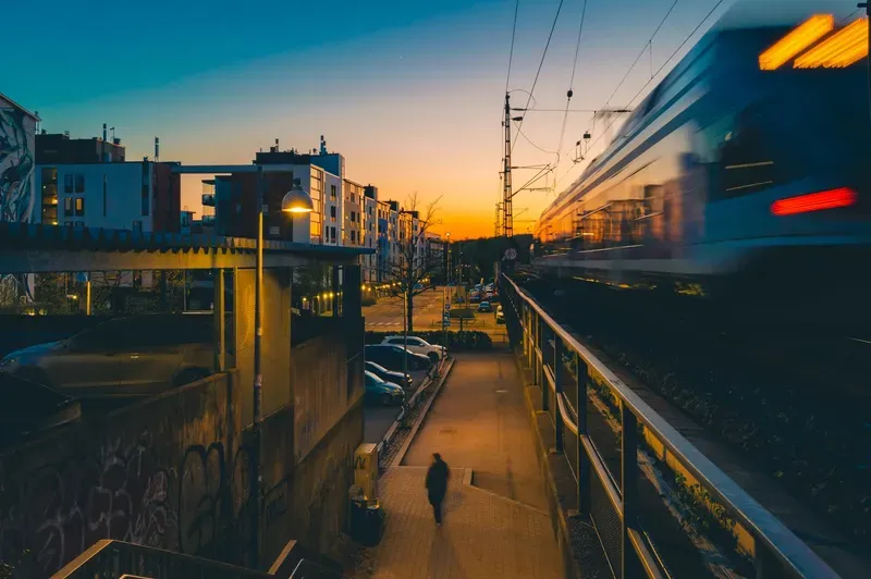 A lone figure walks a footpath beside a graffiti wall as a train blurs past on elevated tracks under a deep orange dusk sky.