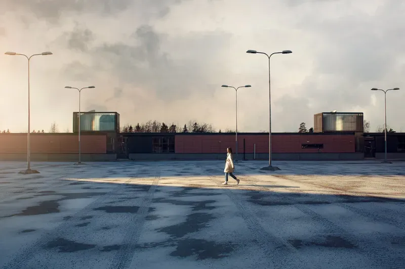 A woman walks alone across a vast empty parking lot at dusk, streetlights standing over her beneath a hazy, glowing sky.