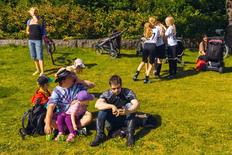 Families on a sunny lawn: a man in a tie-dye shirt with a small girl in a purple cap on his lap, another man in biker boots looking at his phone, and girls in white football shirts hugging beside parked strollers.