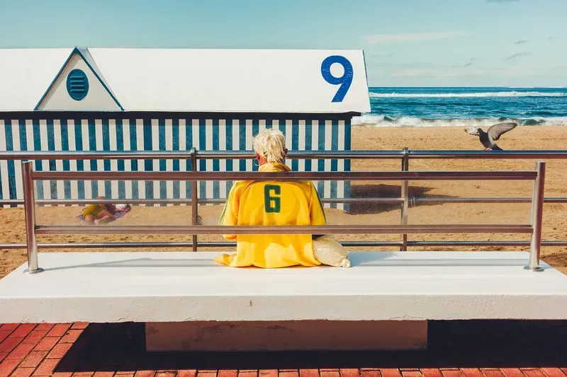 An older woman in a yellow number-six jersey sits on a bench facing a striped beach hut marked nine, a gull flying past on the sand.