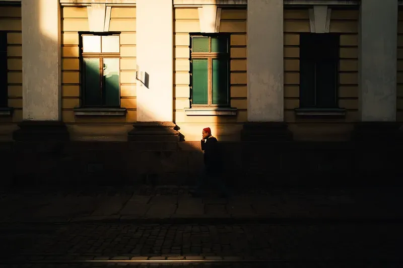 A man in a red beanie walks past a sunlit building facade with green-shuttered windows, caught in a narrow beam of late light.