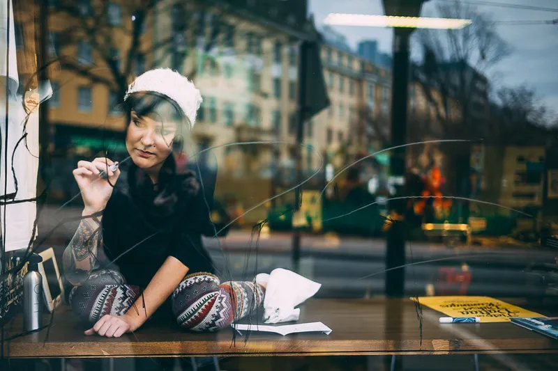 A woman in a white knit hat writes on a café window from inside, the street and buildings outside reflected across the glass.