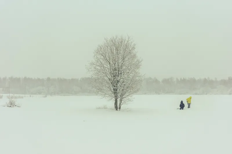 Two children in yellow and navy coats play beside a bare tree in a snow-covered field during a Helsinki snowfall.