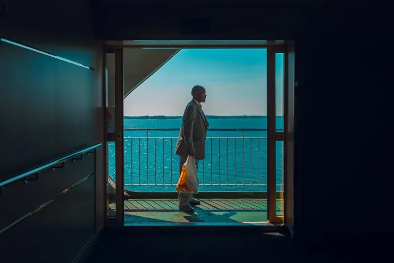 A man carrying a shopping bag steps onto a sunlit ferry deck, silhouetted against the open sea framed by a dark doorway.