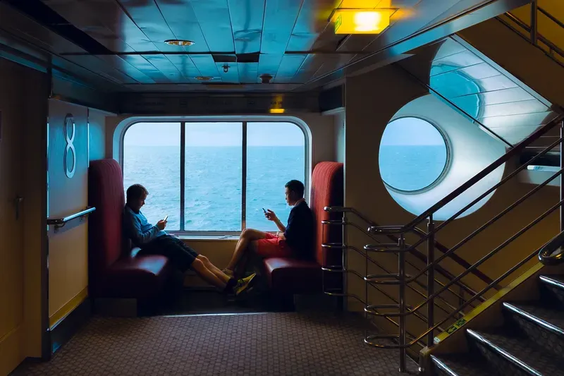 Two teenagers sit on red ferry benches by a panoramic window, absorbed in their phones as the open sea passes behind them.