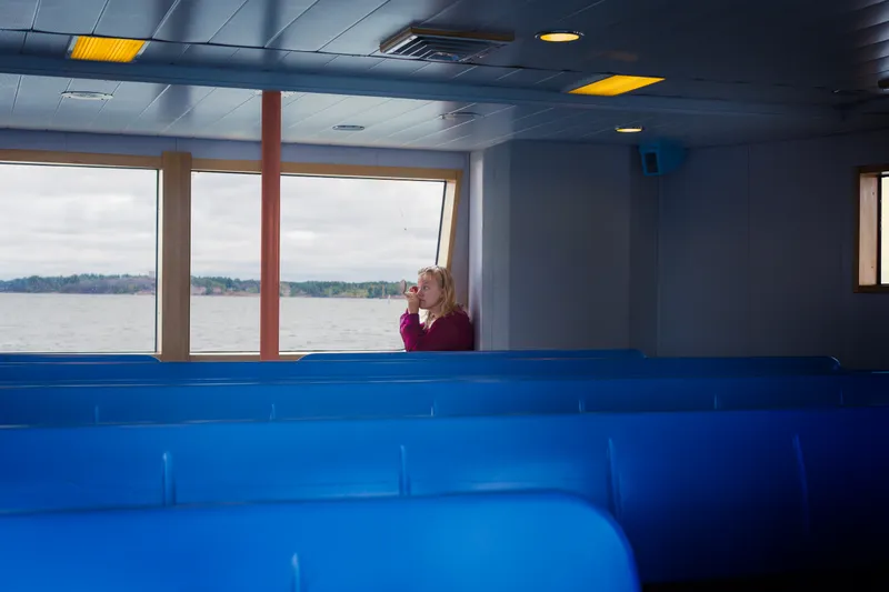 A woman checks a small mirror while seated by a ferry window, rows of empty blue seats stretching in front of her.